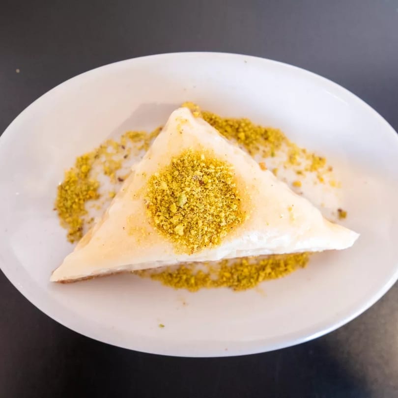 A triangular piece of white dessert, possibly baklava, covered in crushed pistachios on a white plate.