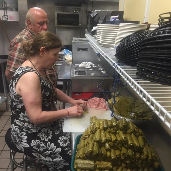 Two people preparing Mediterranean dolmas in a commercial kitchen with stacks of plates.
