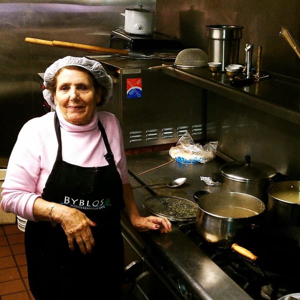 A smiling woman in a hairnet and a Byblos Mediterranean Cafe apron stands in a commercial kitchen.
