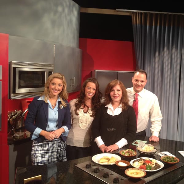 Four people, including a Byblos chef, stand behind a counter with Mediterranean dishes on a TV set.