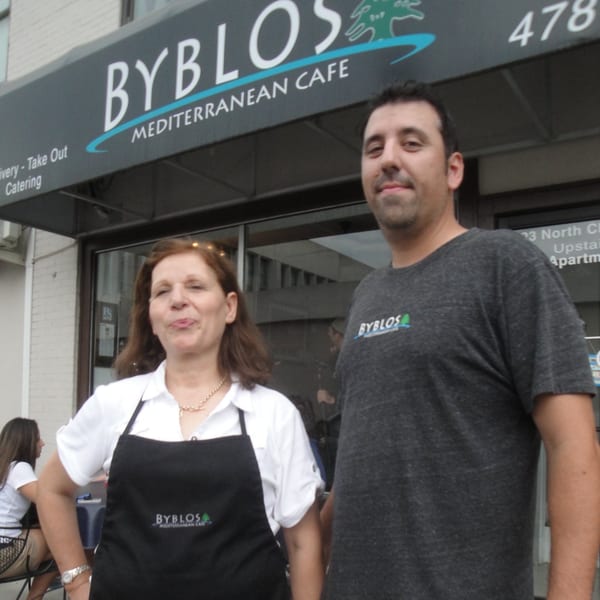 Two Byblos Mediterranean Cafe employees, a woman in an apron and a man in a t-shirt, stand outside the restaurant.
