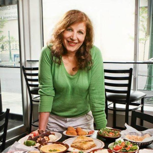 A smiling woman stands behind a table laden with various Mediterranean dishes at Byblos Cafe.