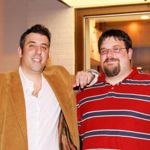 Two men, one in a brown jacket and one in a striped shirt, smiling at the camera.