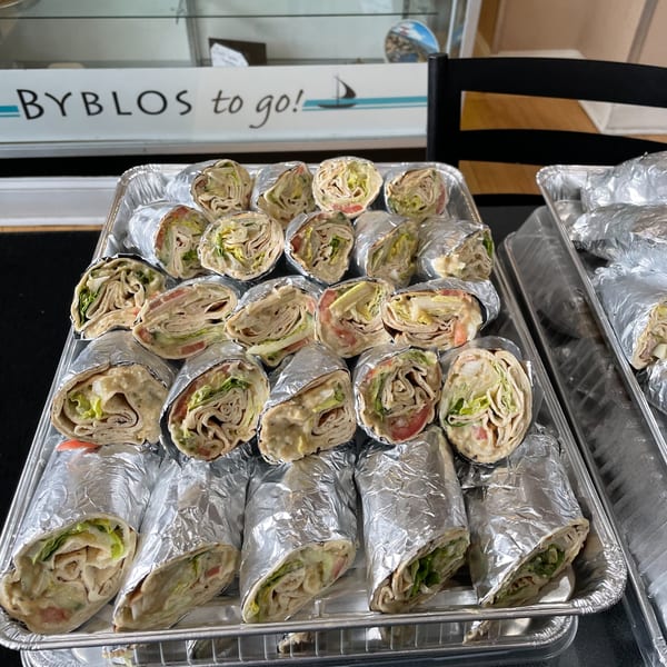 Byblos to Go! sign above two foil trays filled with many hummus and vegetable wraps.