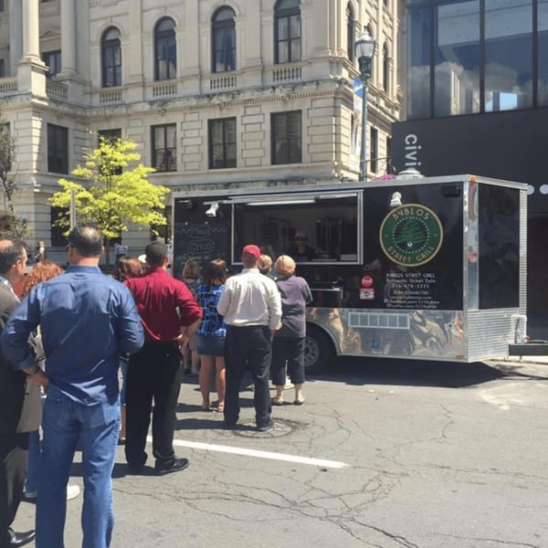 People queueing at the Byblos Street Grill food truck on a sunny day in front of a building.